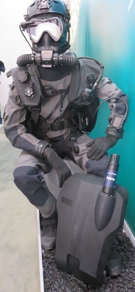 The MCM100 with DiveTrack transponder. The transponder slots into the recess in the back cover of the rebreather, which is shown separate from the equipment. Note the wrist monitor and the LED head-up display in front of the mask.