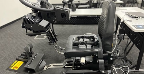 The XR Training consortium's USMC Amphibious Combat Vehicle driver simulator in its seat configuration with two DoF motion platform. The Vive HTC Elite headset is on the seat. The Instructor station laptop is on the table behind.