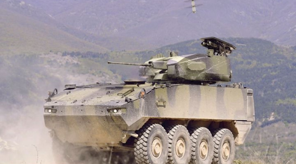 A Piranha 5 8x8 fitted with Rafael's Samson Integrated RWS armed with a 30 mm cannon, pod of two LR2 ATGWs, and a Spike Firefly UAV hovering above.